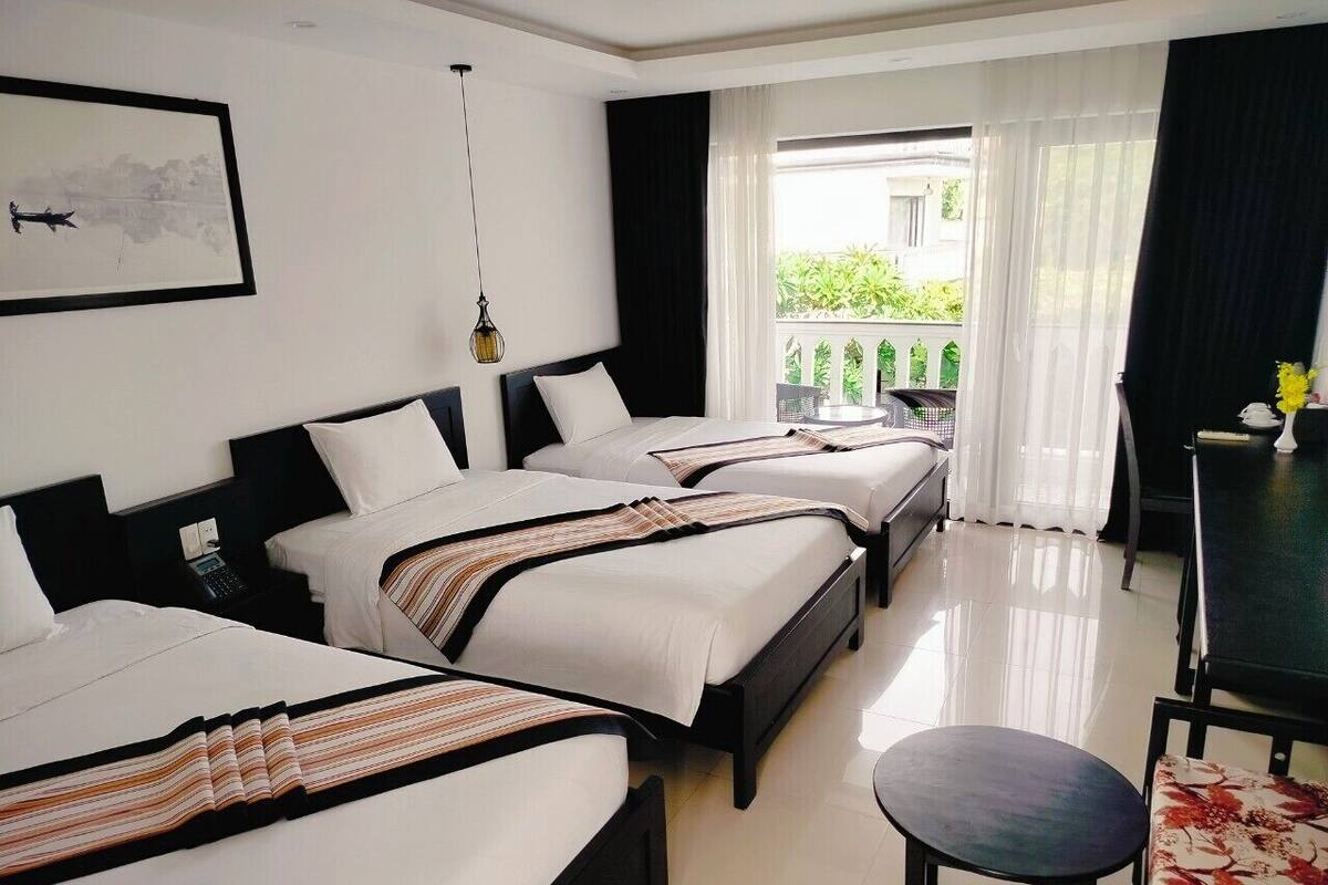 A hotel room with three beds, a dining table, and a view of greenery outside.