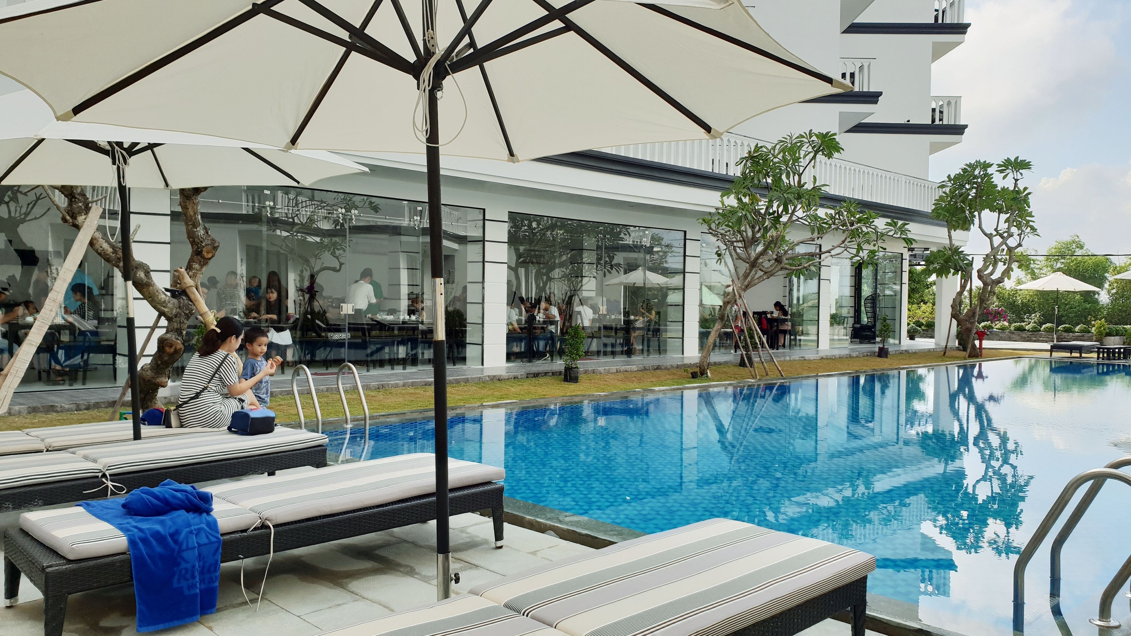 A poolside area with lounge chairs and umbrellas, a glass building with people inside, and a clear blue pool.