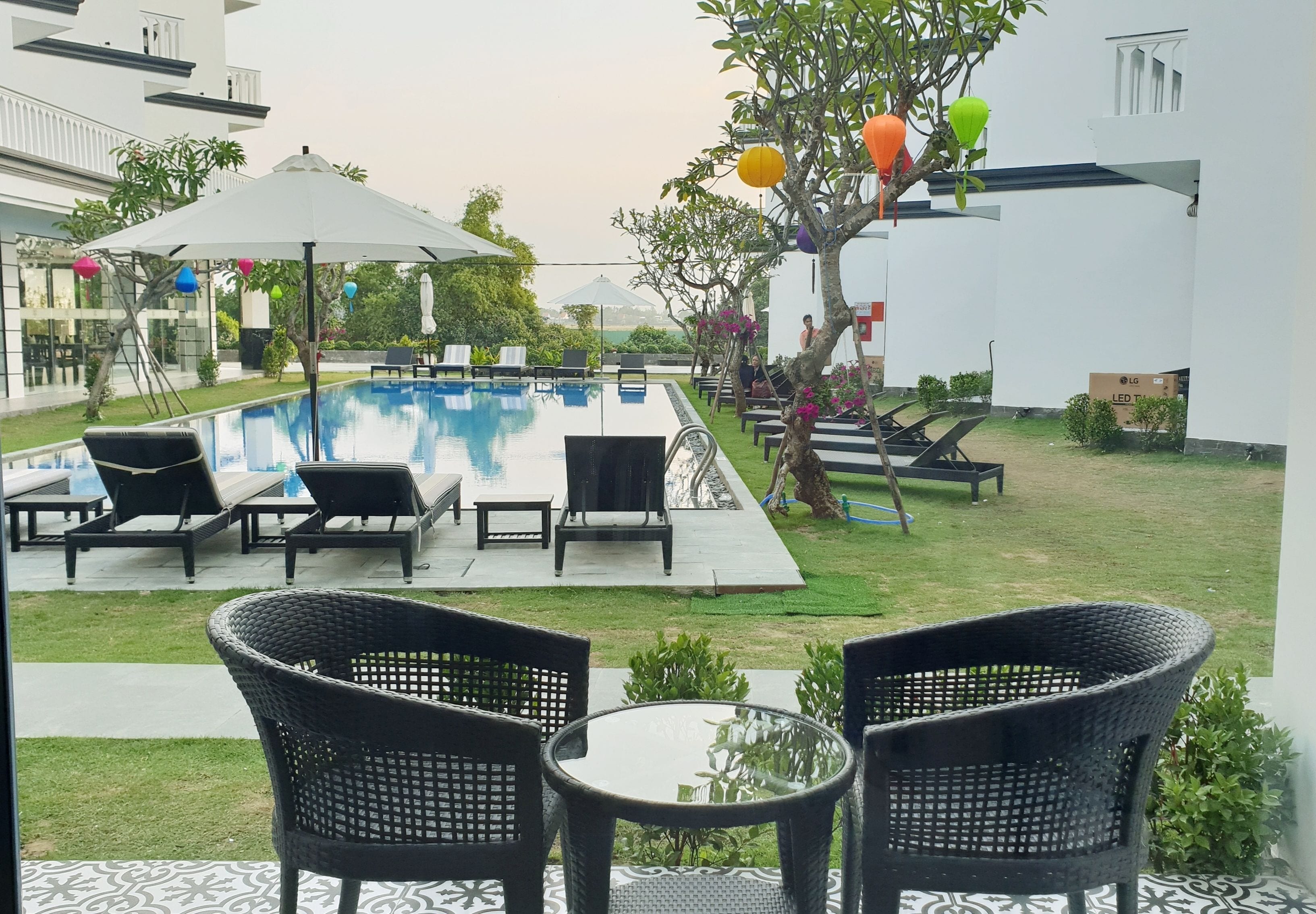 A pool area with lounge chairs and umbrellas, surrounded by greenery and a white building.