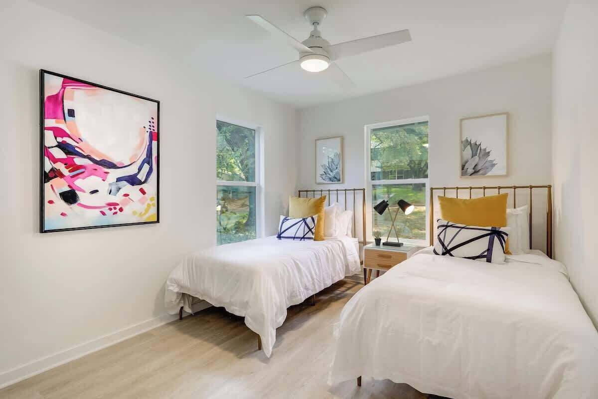 A bedroom with two beds, large windows, ceiling fan, and artwork on the walls.