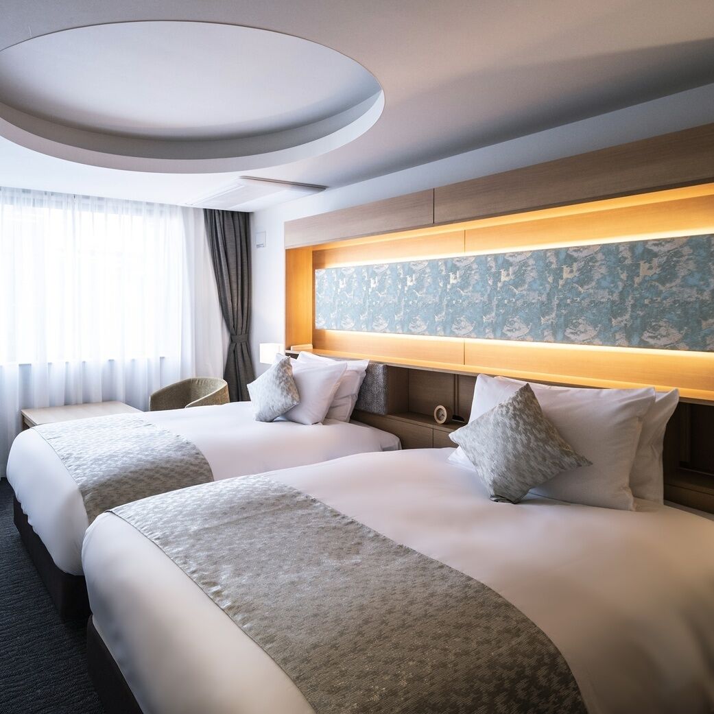 A hotel room with two beds, a large window with curtains, a headboard with a lighted panel, and a round ceiling light.