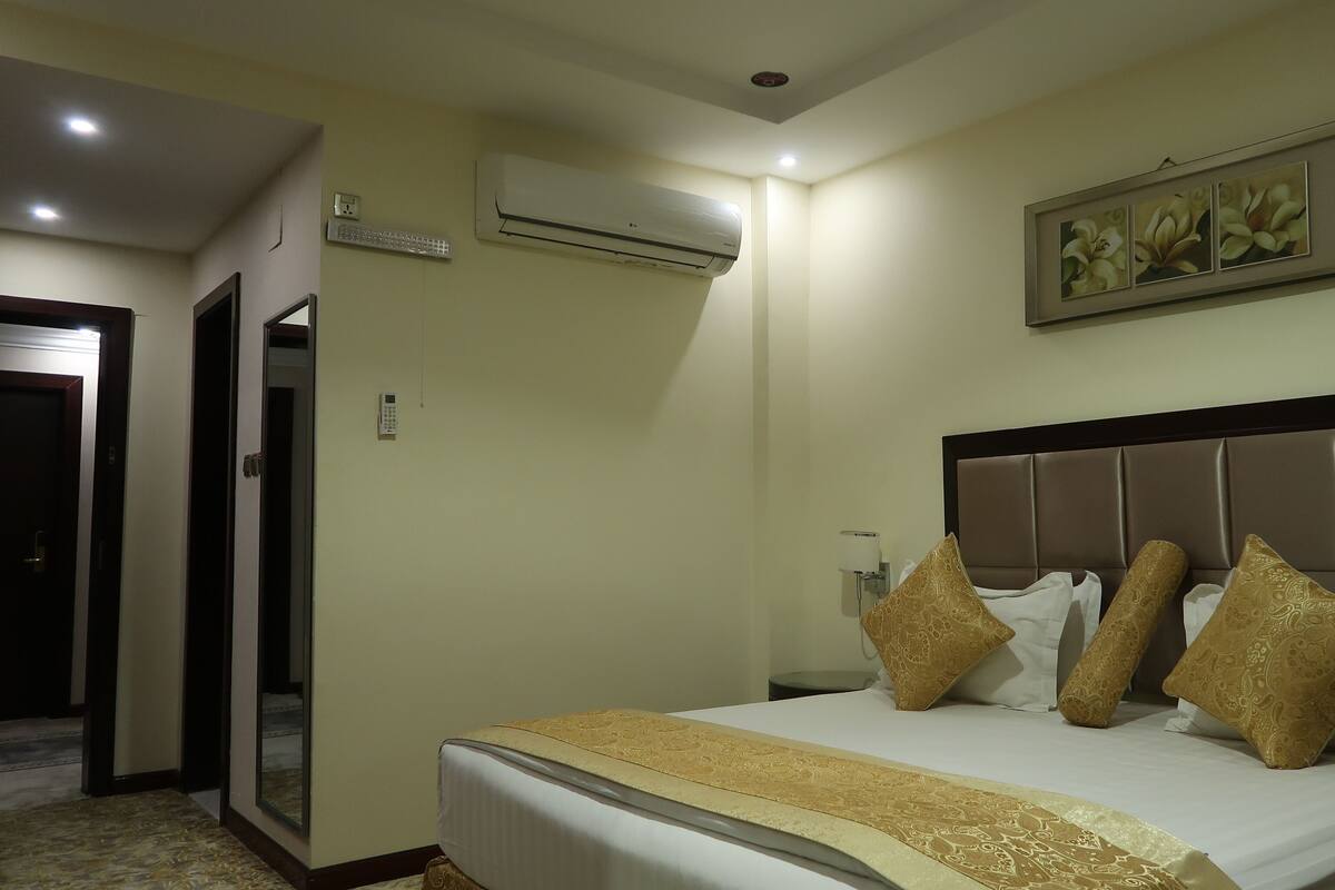 A hotel room with a bed, a wall-mounted air conditioner, a painting, and a door.
