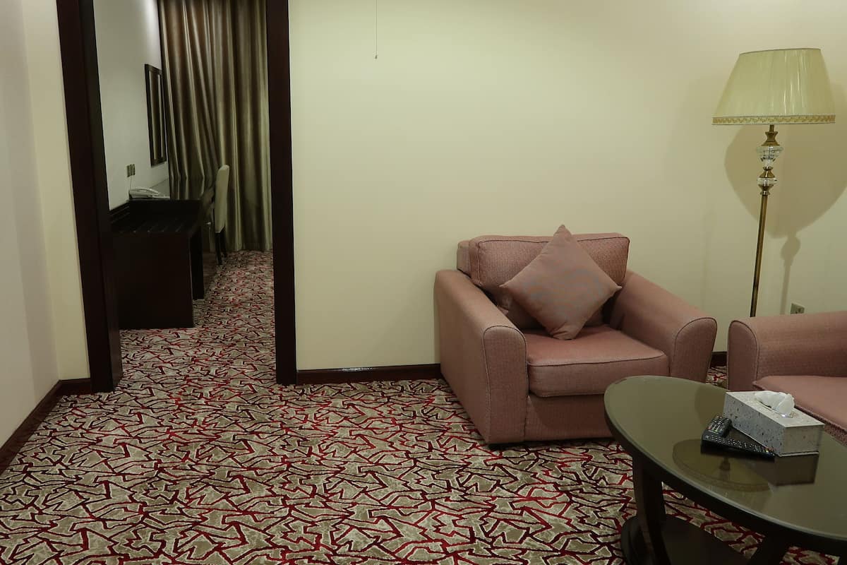 A hotel room with a patterned carpet, a sofa with a pillow, a small table with a remote control and tissues, and a lamp.