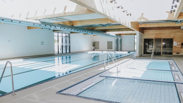 Ein großer, überdachter Schwimmbereich mit mehreren Bahnen, Holzbalkendecke und Glastür-Eingang.