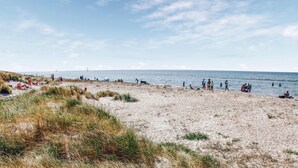 Ein Strand mit badenden und sonnenbächelnden Personen, ein Segelboot in der Ferne und ein klarer Himmel.