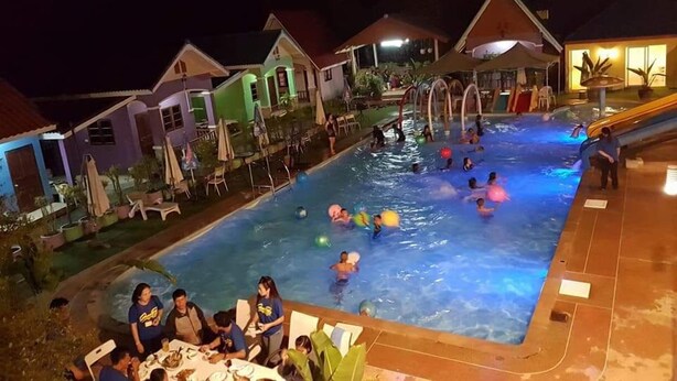 Un espace piscine où des personnes nagent et s’amusent, entouré de maisons dotées de coins salon extérieurs.