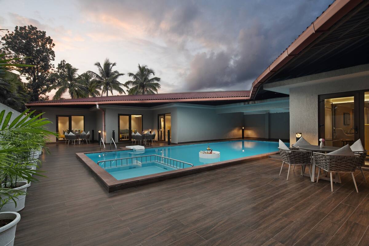 Una piscina con terraza de madera, mobiliario exterior y palmeras al fondo.