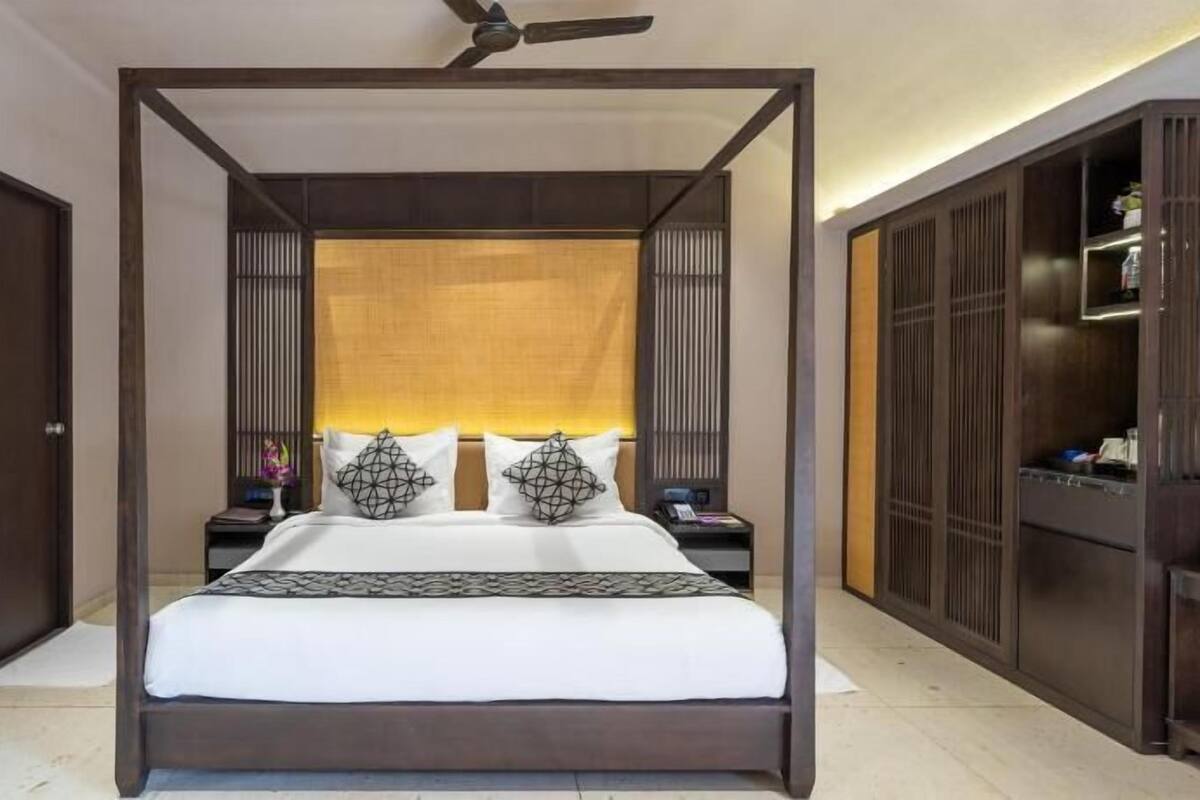 Una cama con dosel y estructura de madera, un ventilador de techo y una mesita de noche con una lámpara y un florero.