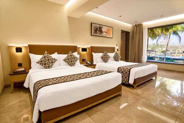 Two double beds with geometric-patterned bedspreads and matching pillows, a nightstand, and a table with a view of palm trees and a pool.