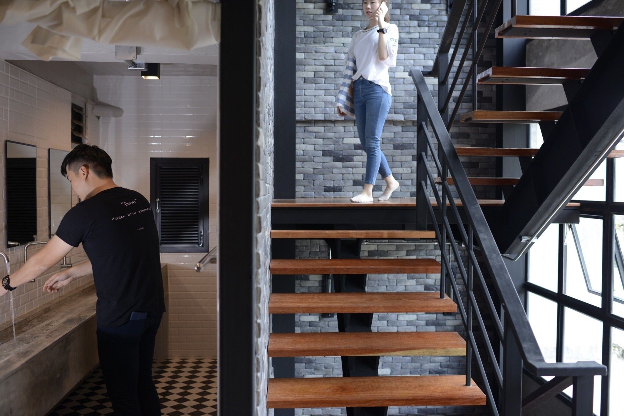 Un intérieur moderne avec un sol à carreaux, un escalier avec des marches en bois et des rampes en métal, et une personne utilisant un lavabo.