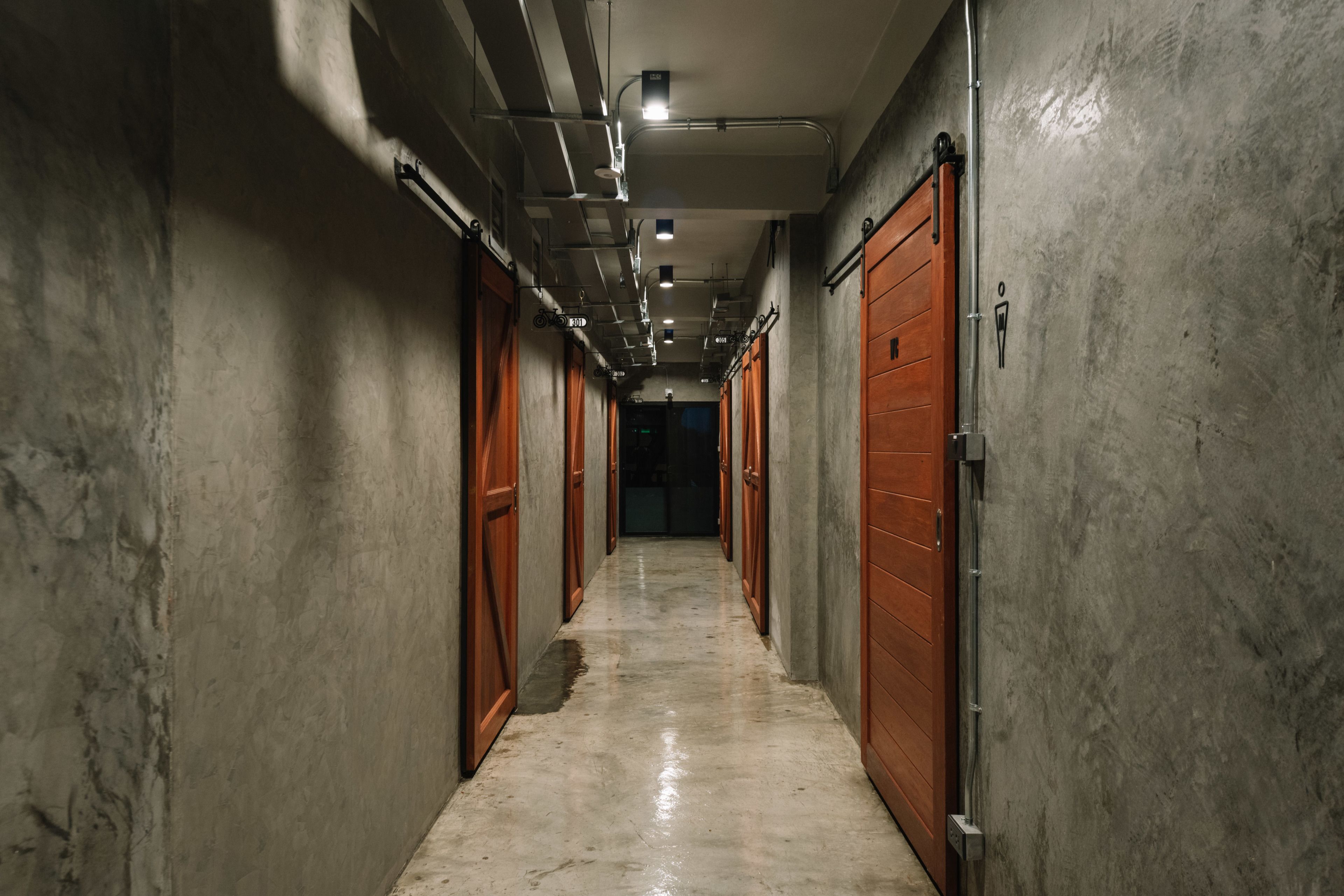 A long, narrow hallway with concrete walls and wooden doors, leading to a darkened area at the end.