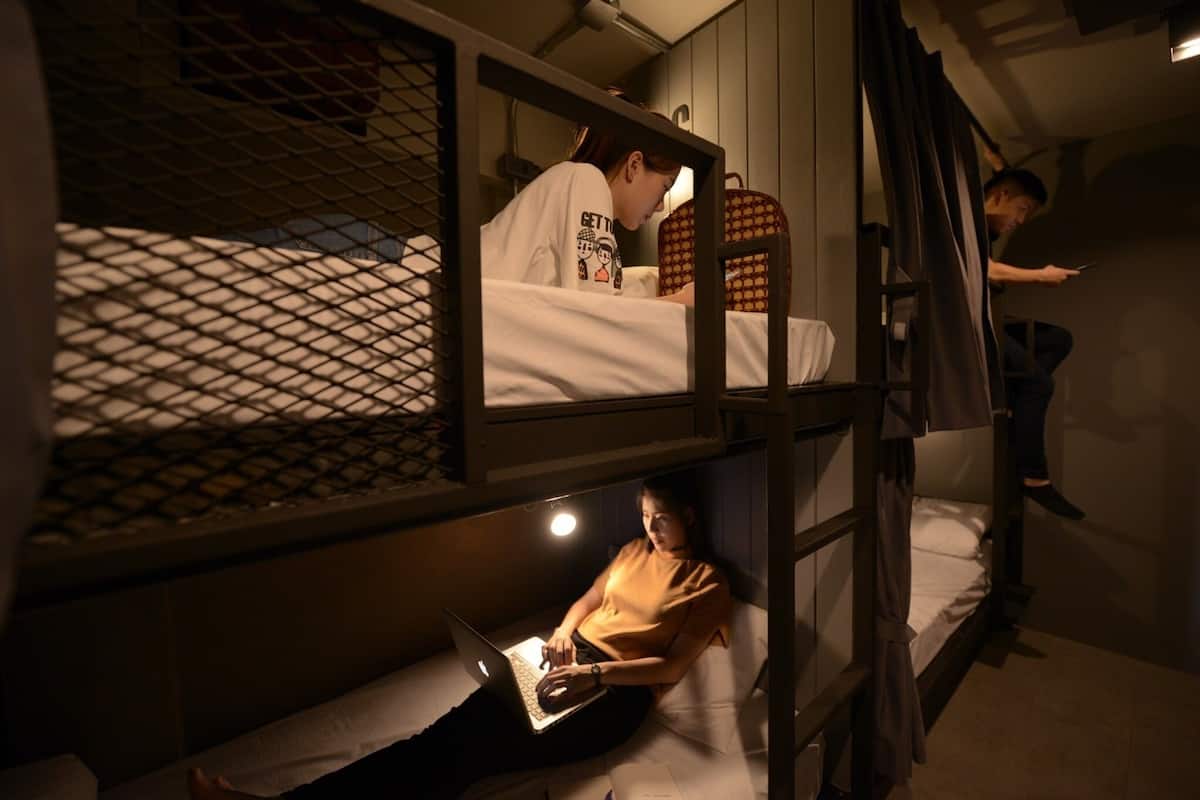 Quarto em dormitório com beliches, um laptop no beliche de baixo e uma pessoa usando um dispositivo móvel.