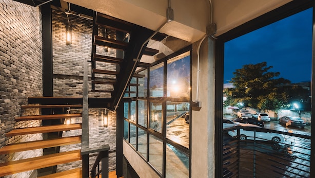 A modern interior with a wooden staircase, a brick wall, and large windows overlooking a parking area. - Bed & Bike Hostel (Chiang Rai)