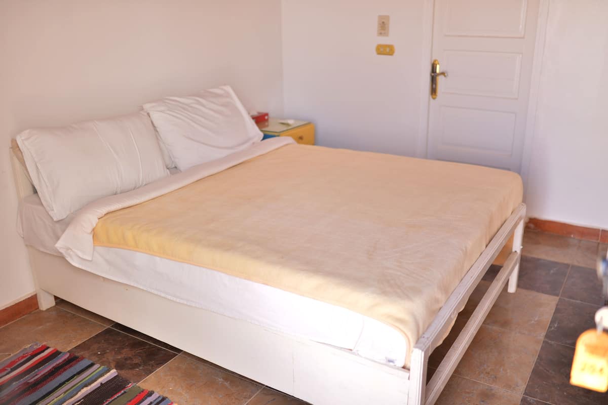 Ein Bett mit weißer Bettwäsche und einer beige farbenen Decke, ein Nachttisch mit einer Lampe sowie eine weiße Tür mit goldener Griff.