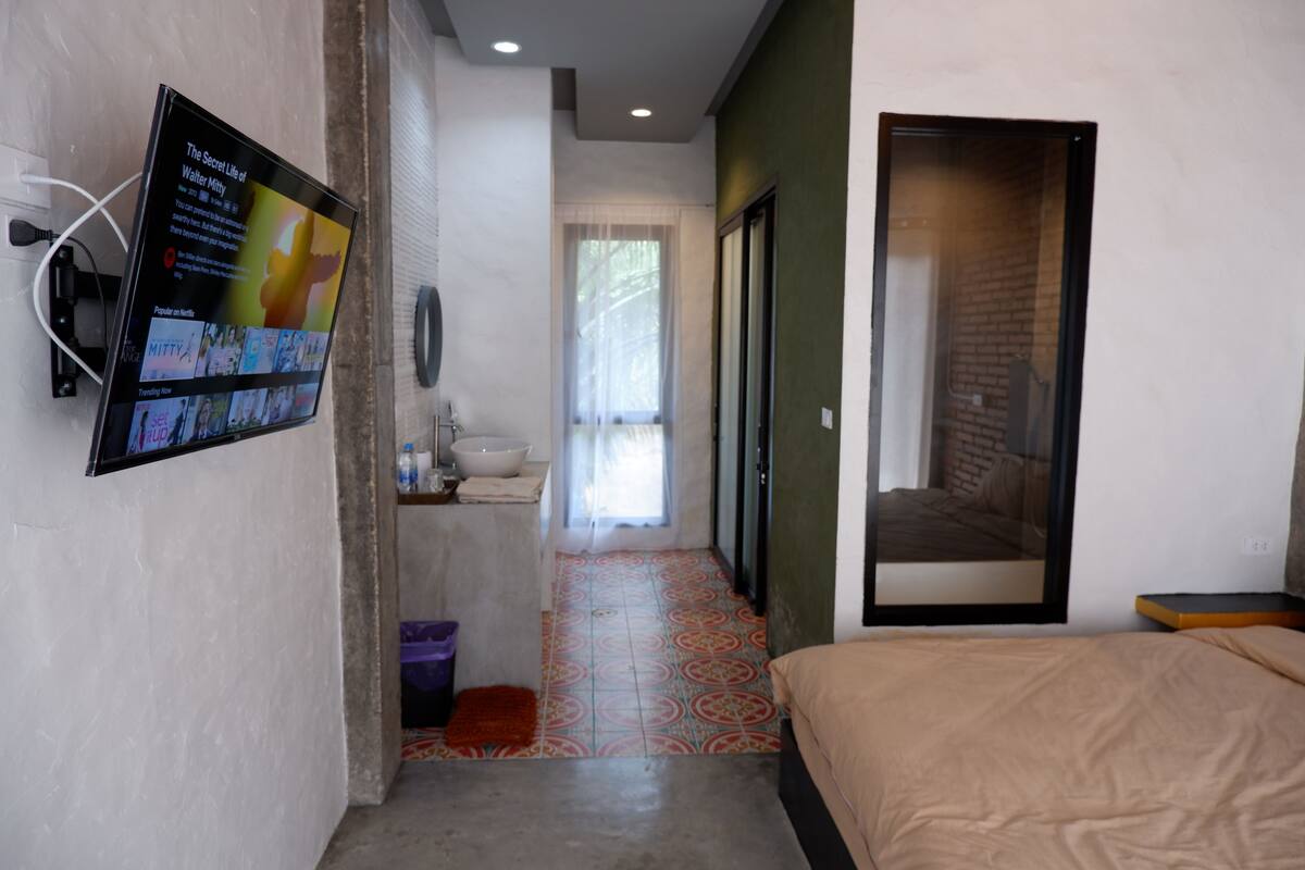 A modern hotel room with a large flat-screen TV mounted on the wall, a bed with a beige cover, a small bathroom with a sink and mirror, and a colorful patterned floor.