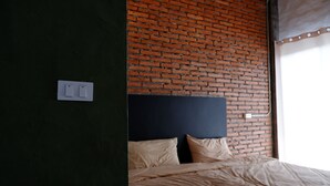 Quarto com parede de tijolos, cama com roupa de cama bege e painel elétrico branco na parede.