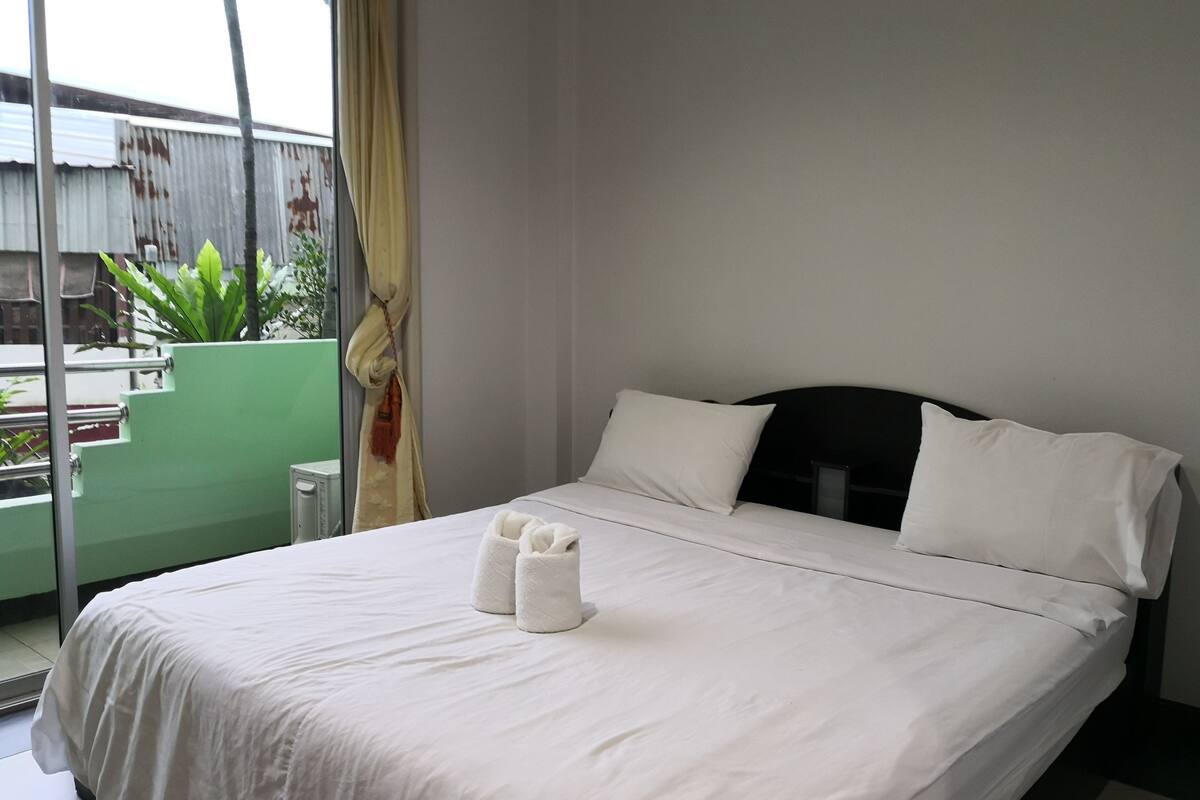 Quarto de hotel com cama, almofadas, janela com cortinas e vista para um edifício.