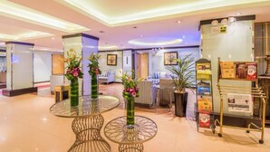 Un hall d’hôtel avec des tables rondes en verre, des plantes en pot et un kiosque en libre-service.