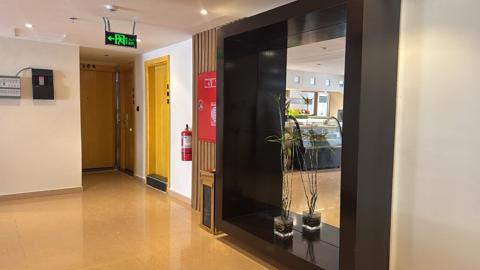 Pasillo de hotel con extintor, señal de salida y plantas en macetas.