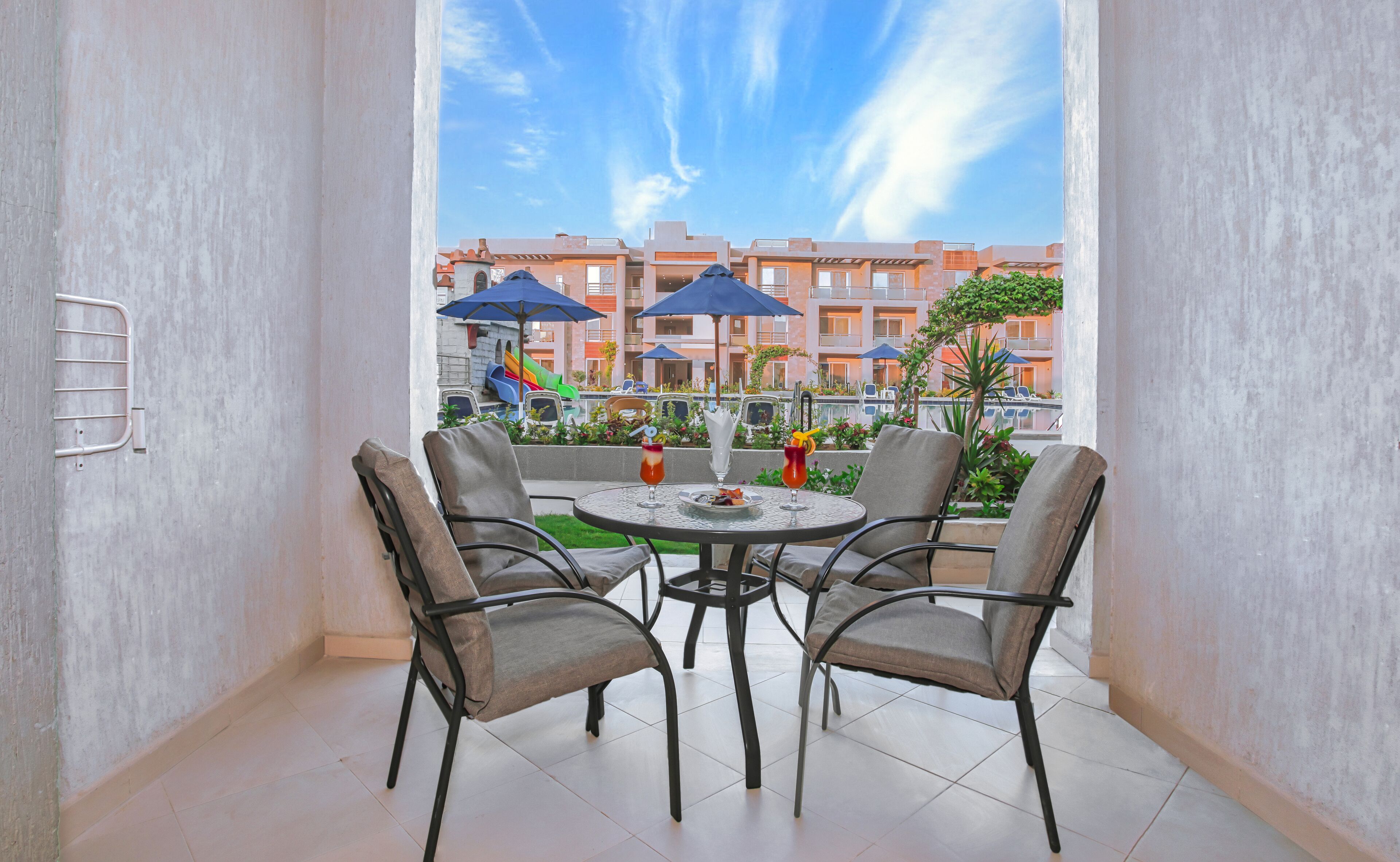 A balcony with a round table and four chairs, overlooking a playground and buildings.