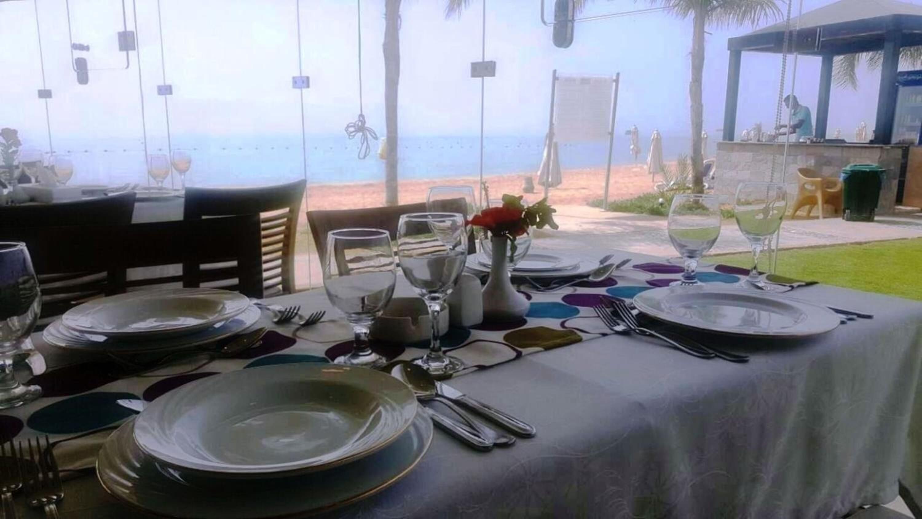 An outdoor dining setup with a view of the beach, tables set with plates, glasses, and cutlery.