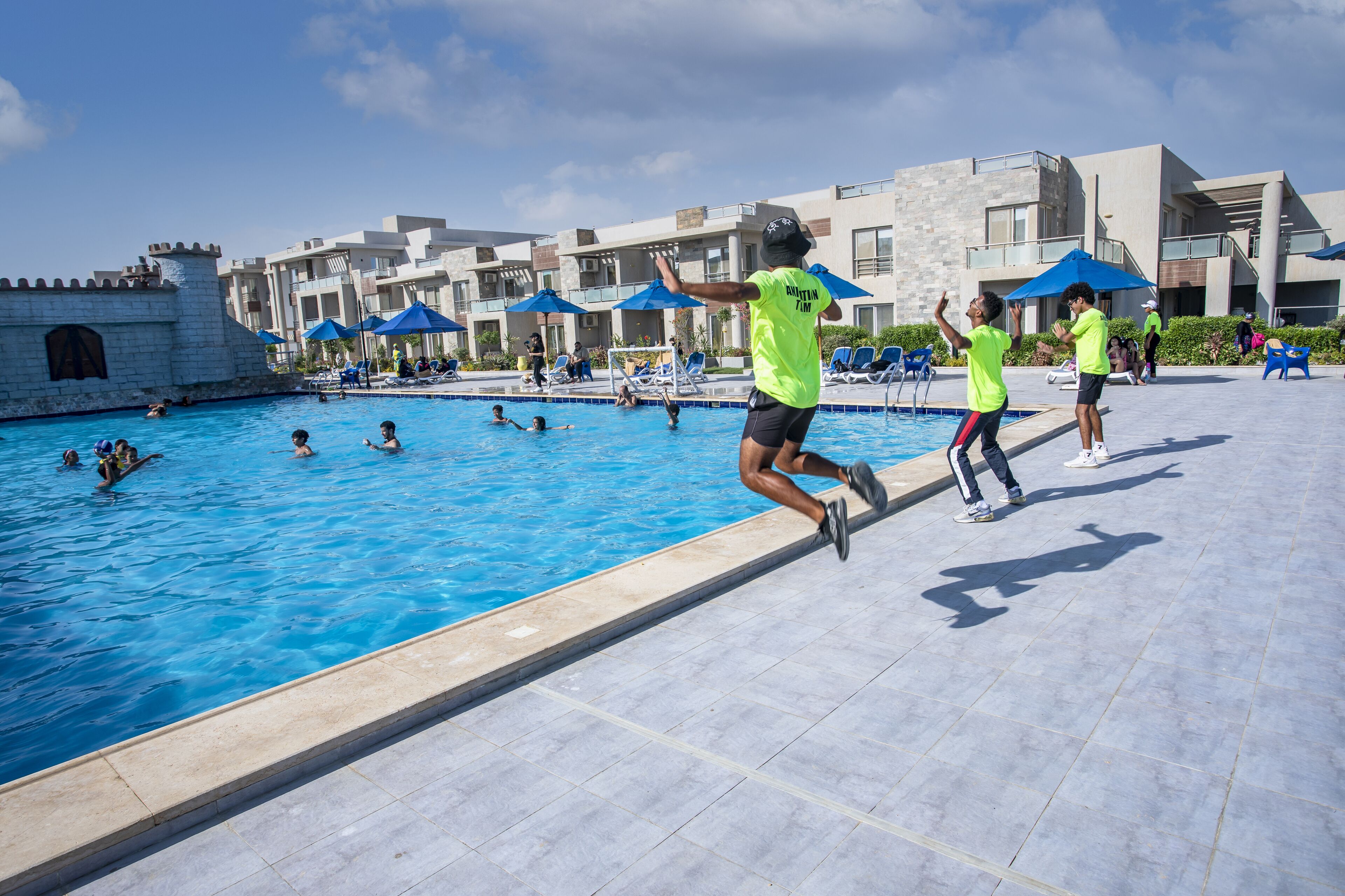 A swimming pool area with people jumping into the water, a castle-like structure, and modern buildings with blue umbrellas.