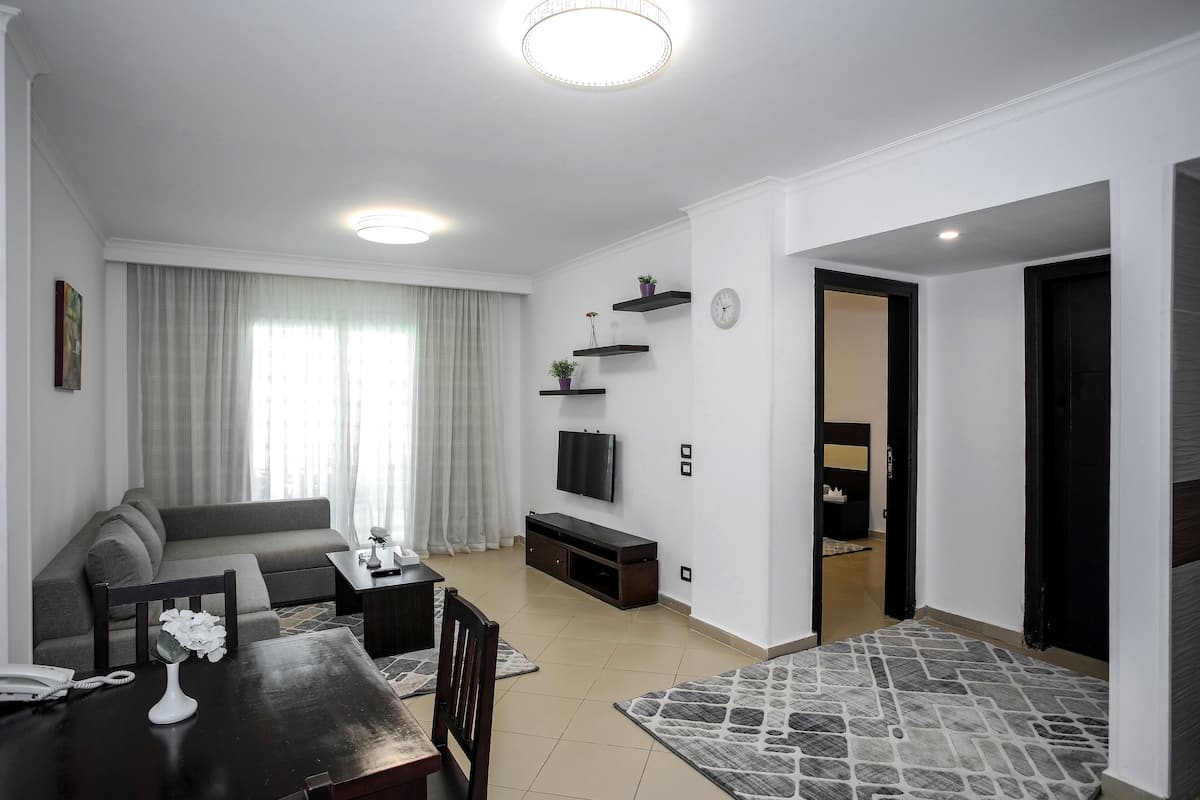 Een moderne woonkamer met een bank, salontafel en televisie. Er staan planten en een klok op de wanden, met boekenplanken.