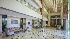 Un hall d’entrée spacieux avec un plafond haut, une colonne centrale et un sol à motifs. On y trouve des coins salon avec des canapés et des fauteuils, ainsi qu’un comptoir de réception à l’arrière-plan.