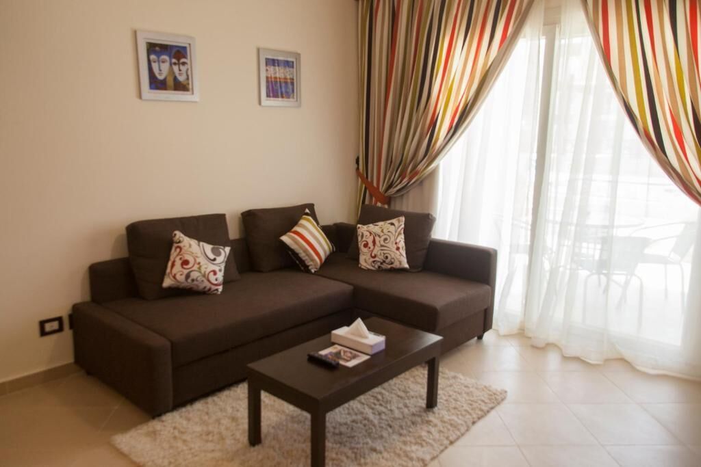 A living room with a brown sofa set, a coffee table, and a white rug. There are two framed pictures on the wall and a window with striped curtains.