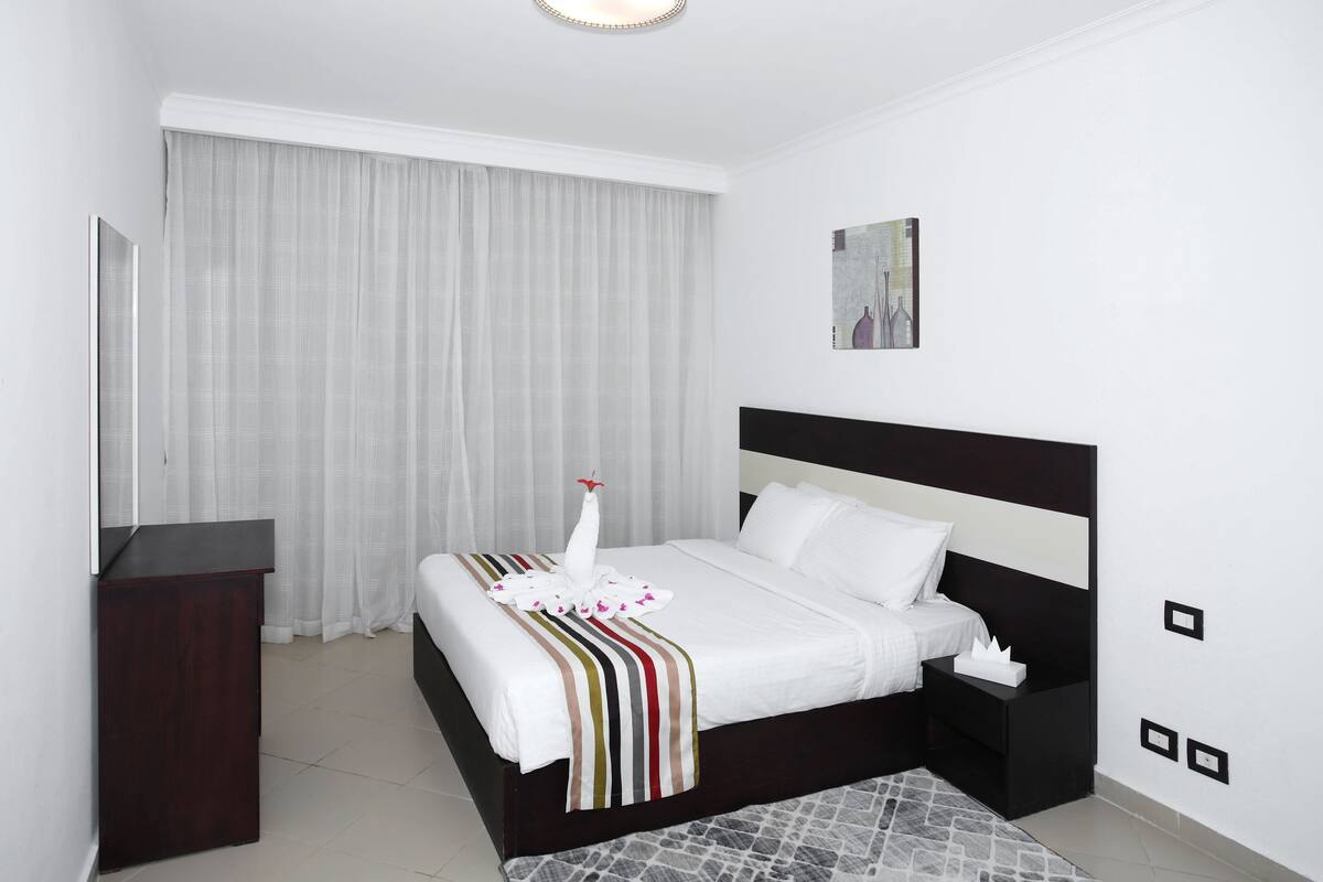 Een moderne hotelkamer met een groot bed, nachtkastje, dressoir, een schilderij aan de muur en een raam met gordijnen.