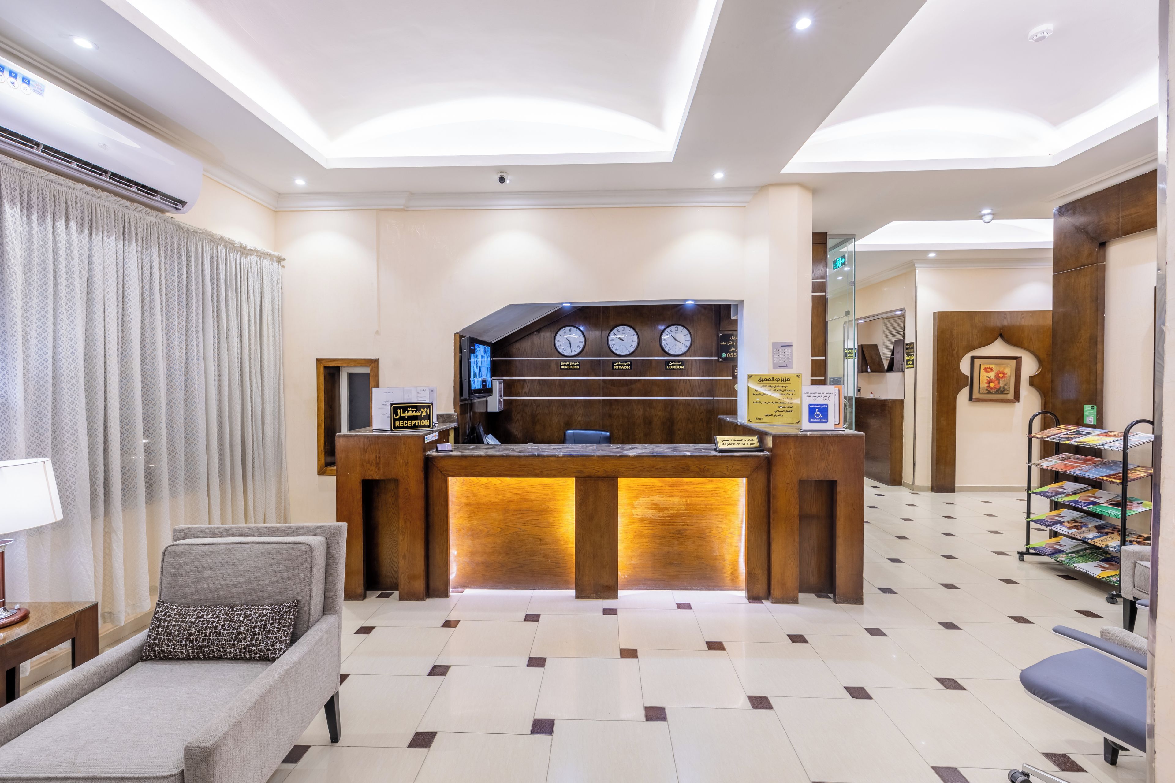 A hotel reception area with a reception desk, a waiting area with a sofa and armchair, and a wall with clocks showing different time zones.