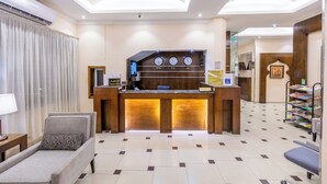 A reception area with a wooden desk, a reception sign, a clock, and a seating area with a chair and a sofa. - Nawara AL  ASKARY - Economic (Riyadh)