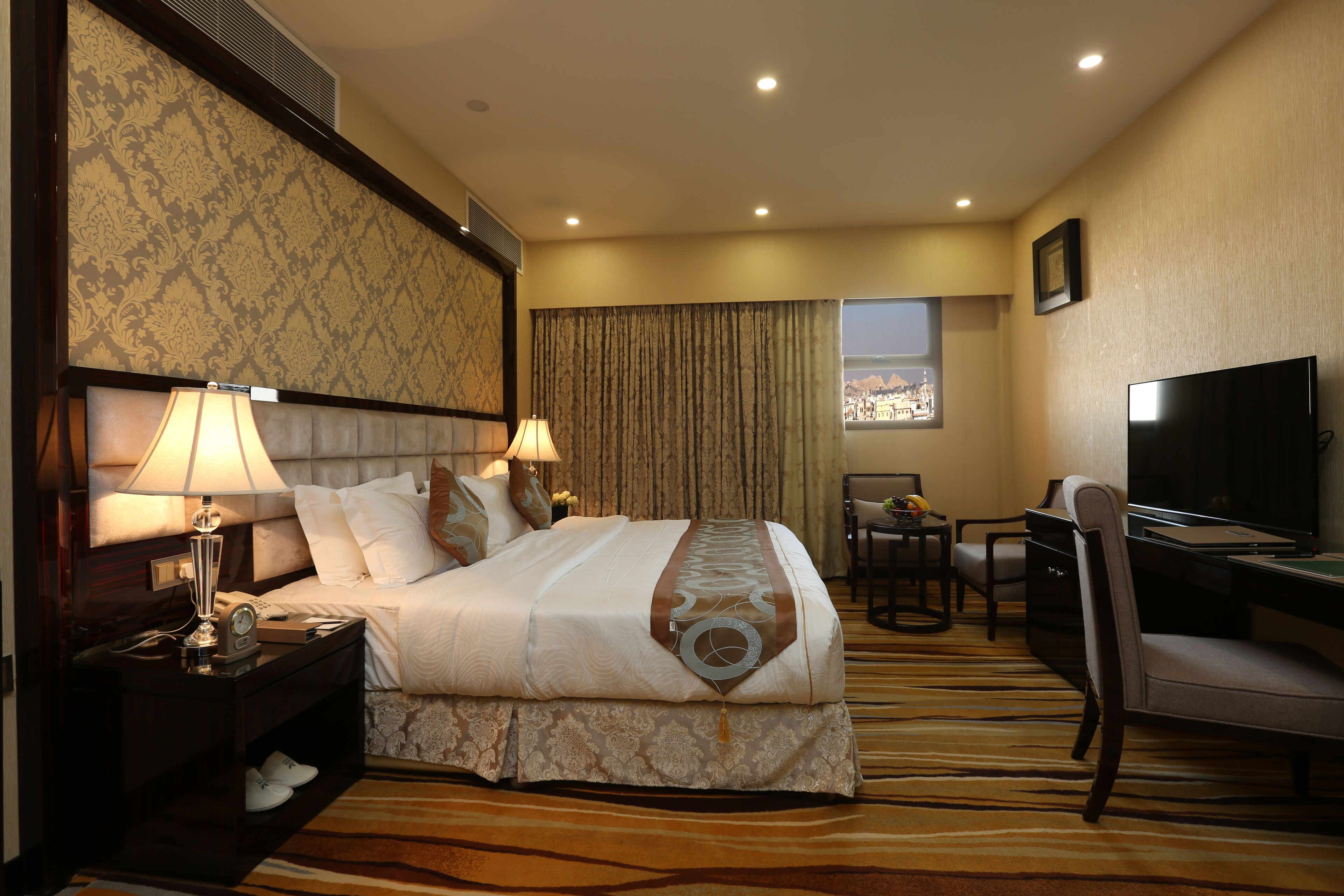 A hotel room with a large bed, a desk with a chair, a television, and a window with a city view.