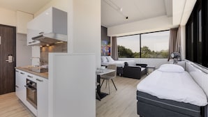 A modern hotel room with a kitchenette, a bed, a desk, and a large window offering a view of the outdoors.