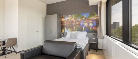 Een moderne hotelkamer met een bed, een bank, een bureau en een groot raam met uitzicht over de stad.