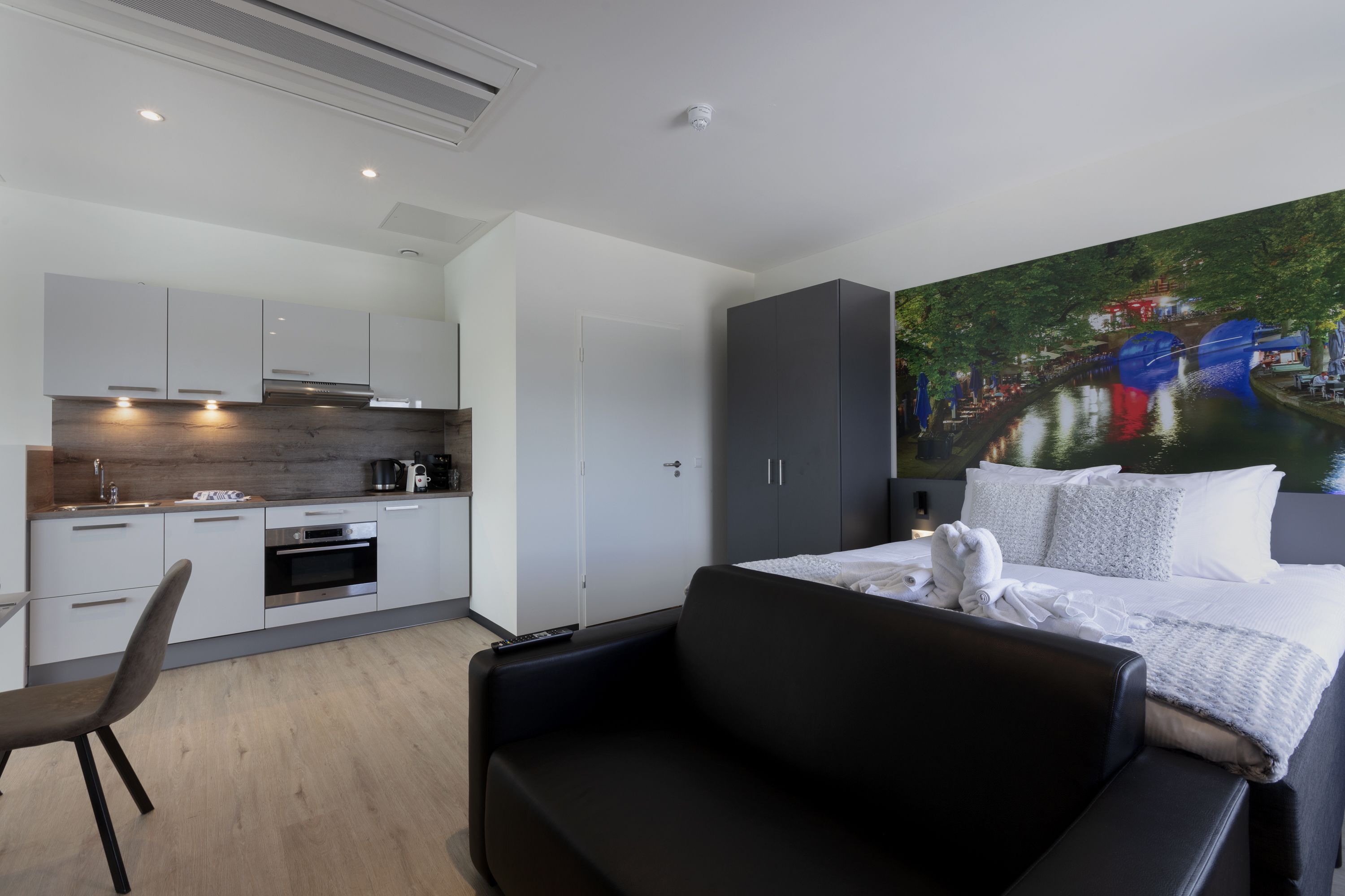 A modern hotel room with a kitchenette, a large bed with white bedding, a black sofa, and a mural of a river scene.