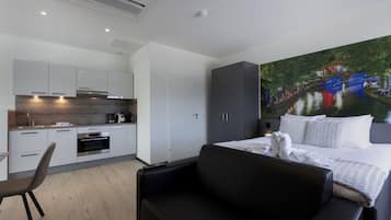 A modern hotel room with a kitchenette, a large bed with white bedding, a black sofa, and a mural of a river scene.