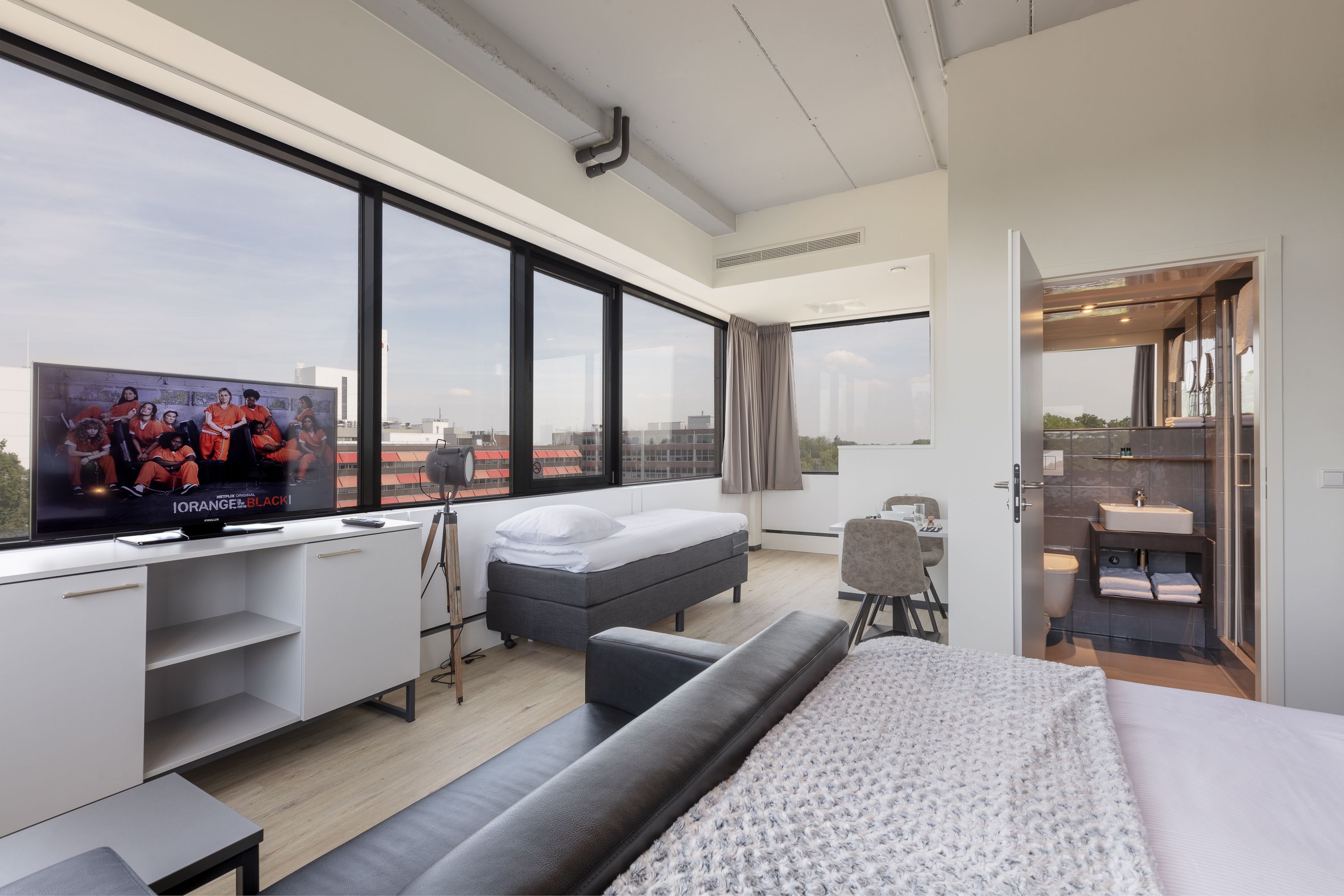 A modern hotel room with a large TV, a bed, a sofa, a desk, and a bathroom.