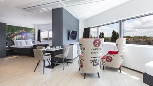 A modern hotel room with a large window, a sofa, a dining area, and two coffee-themed chairs.