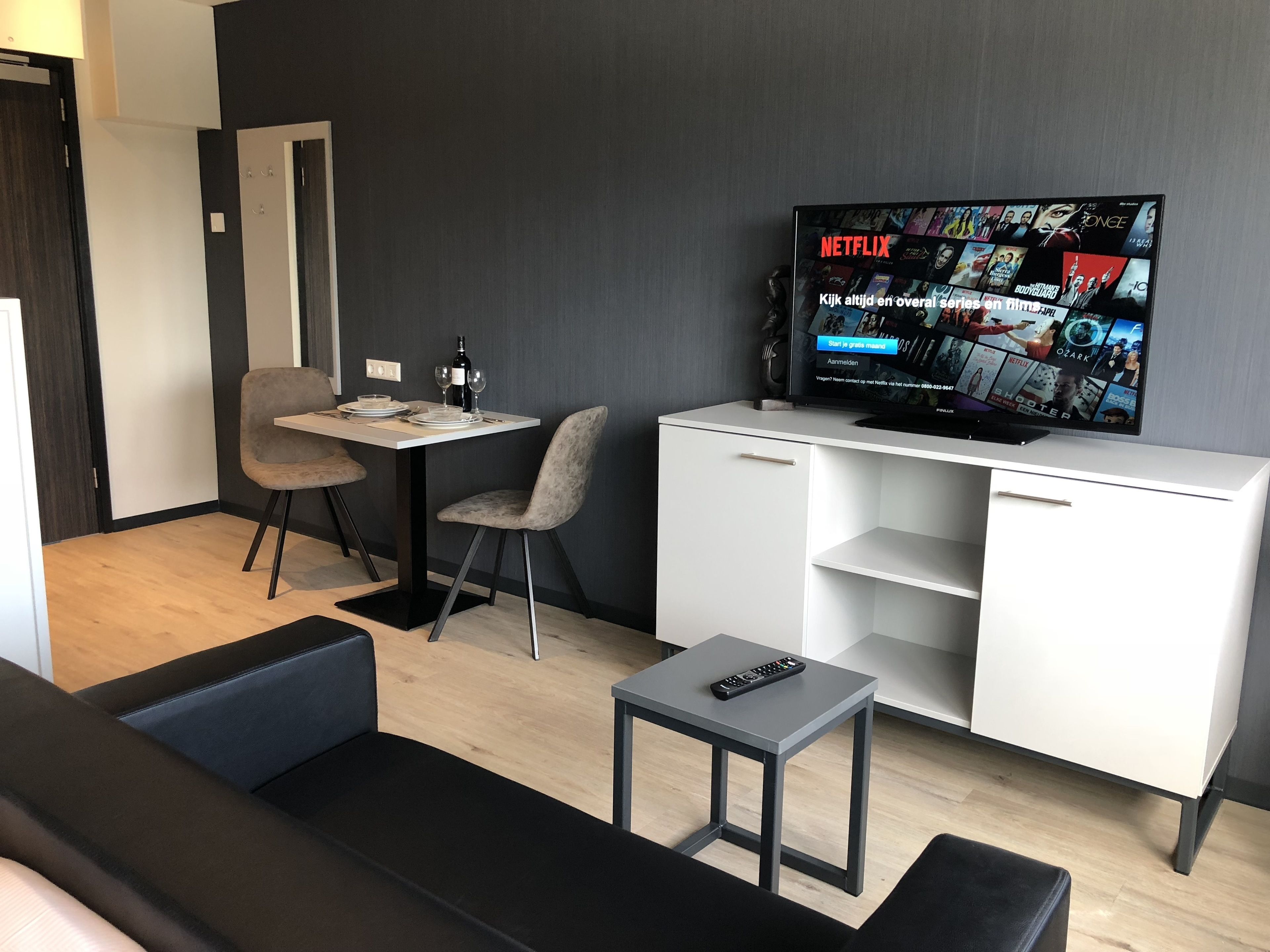 A modern living room with a flat-screen TV displaying Netflix content, a sofa, a small table with a remote control, and a dining area with a table and chairs.
