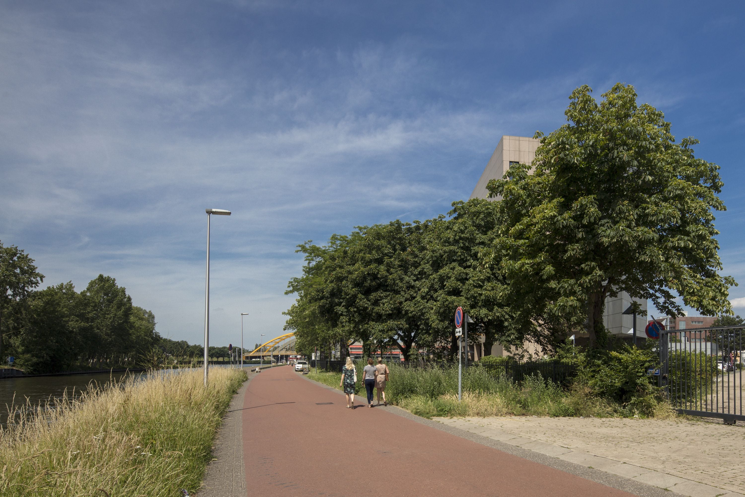 Un sendero pavimentado junto a un canal, con árboles y un edificio al fondo.