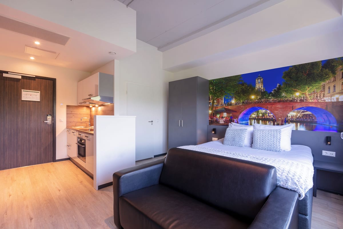 A modern hotel room with a bed, a sofa, a kitchenette, and a mural of a cityscape.