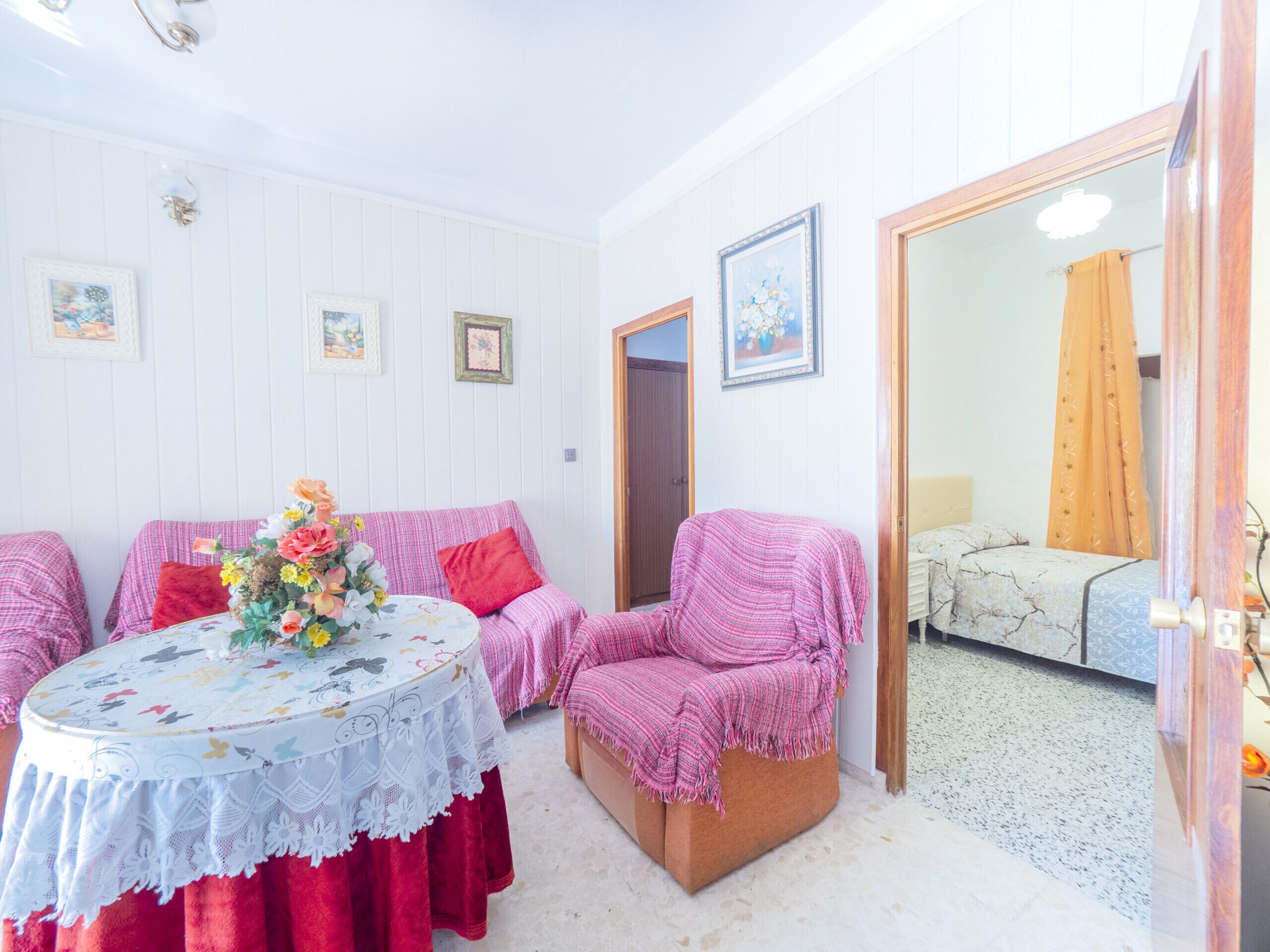 A cozy room with a round table covered with a lace tablecloth, surrounded by two chairs with pink blankets. There are framed pictures on the wall and a wooden door leading to another room.