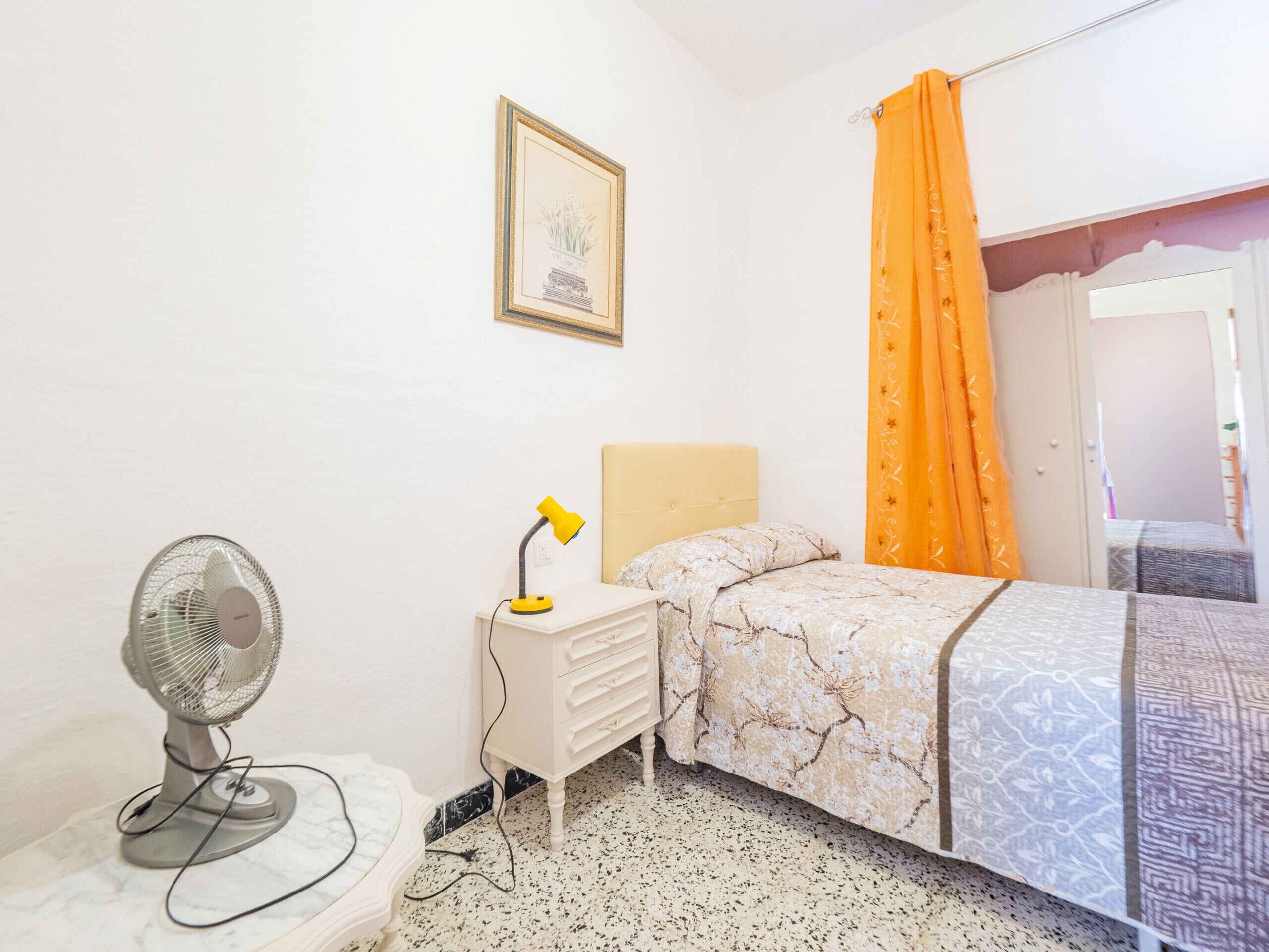 A bedroom with a bed, a bedside table with a lamp, a fan, and a framed picture on the wall.