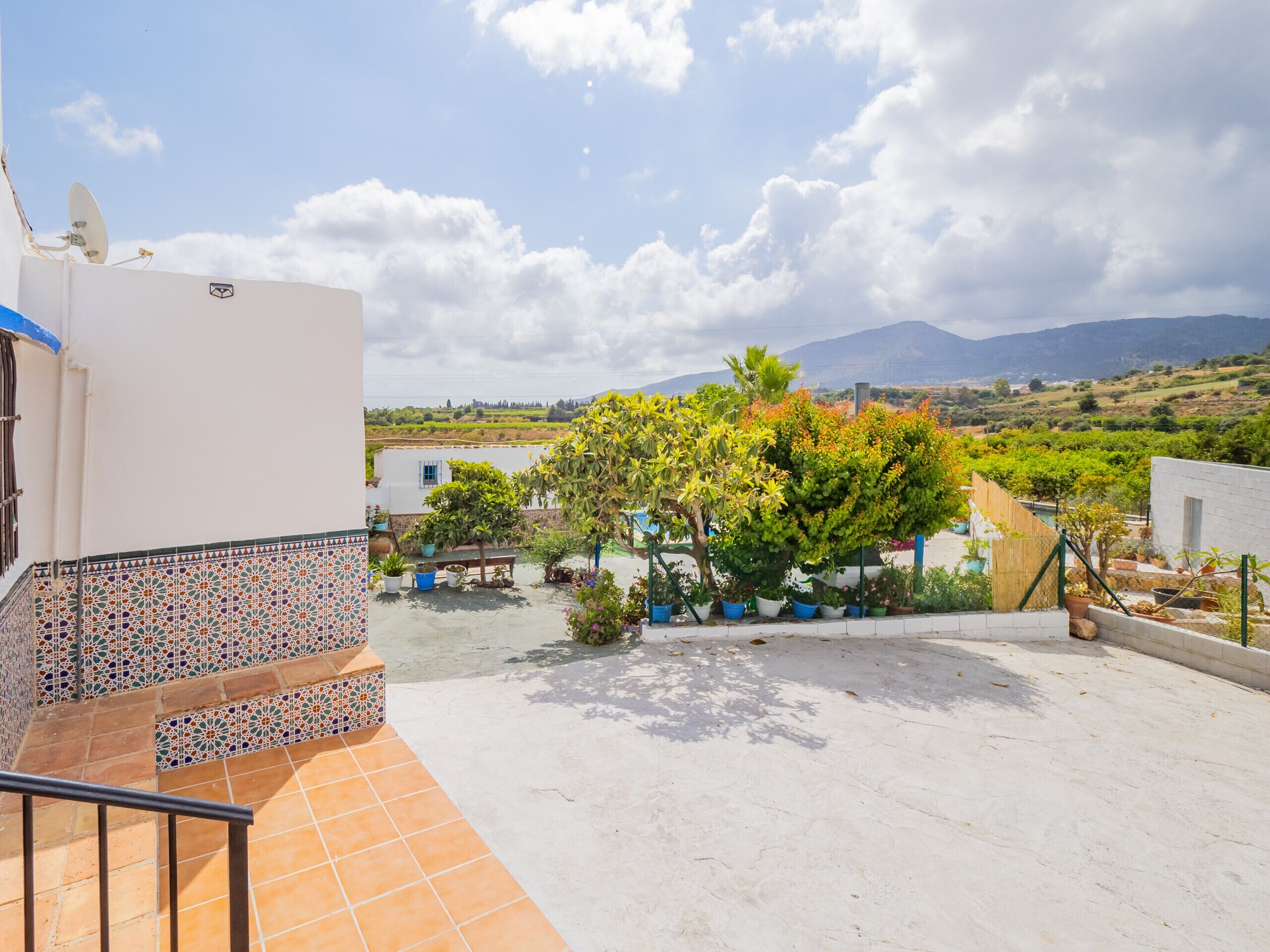 A rooftop terrace with a tiled floor, potted plants, and a view of a rural landscape with mountains in the distance.