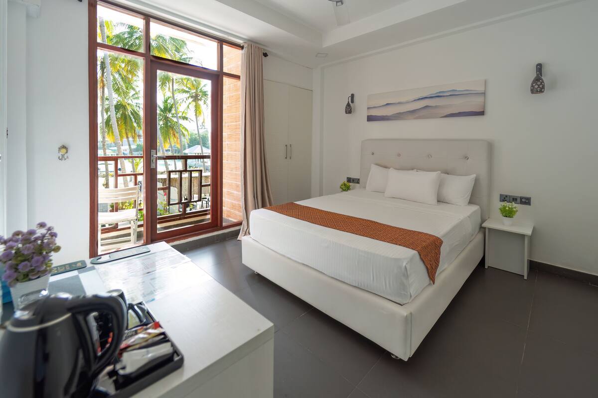 A bedroom with a large bed, a desk with a kettle, and a view of palm trees through the sliding glass door.