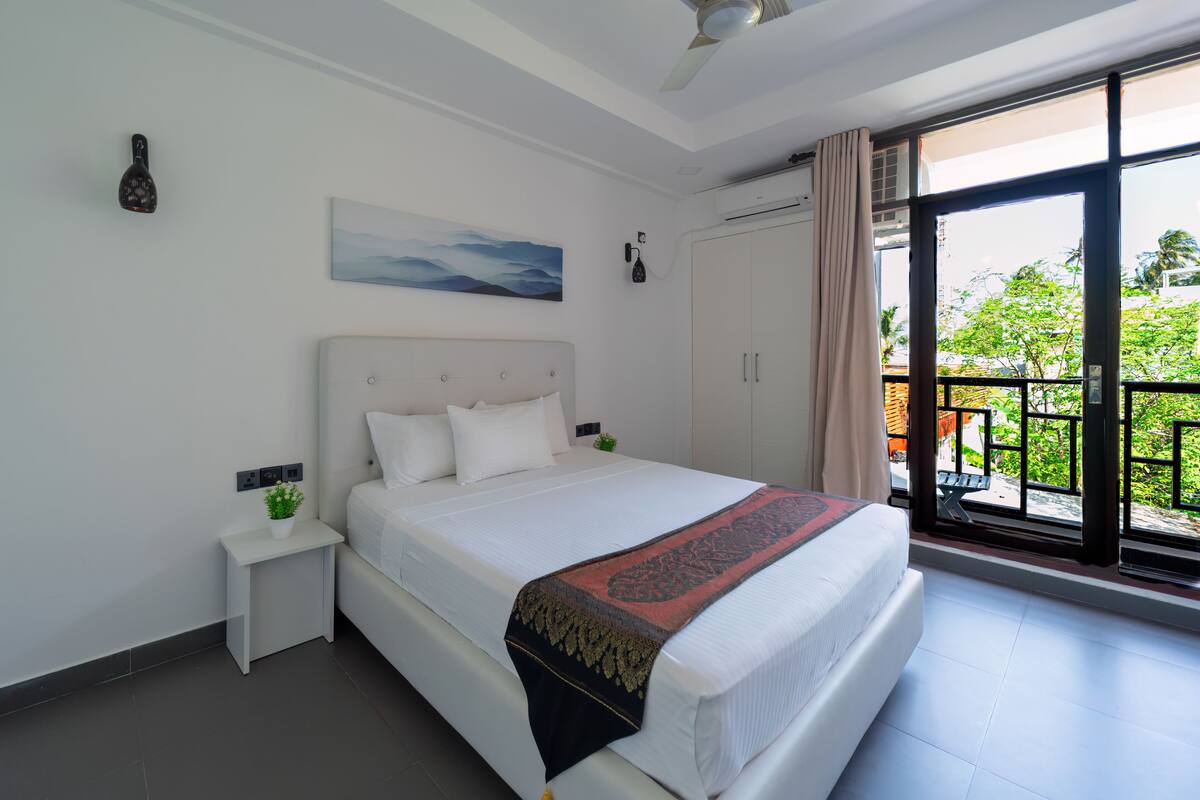 A modern hotel room with a large bed, a small bedside table with a plant, a ceiling fan, and a view of the outdoors through a large window.