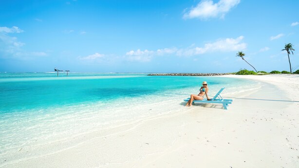 A beach with clear turquoise water, a woman sitting on a blue lounge chair, and palm trees in the background. - Silver Oasis Maldives (Huraa)