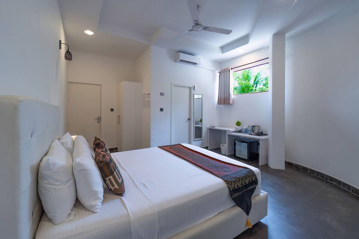 A hotel room with a bed, a desk, a mirror, and a window with a view of greenery.