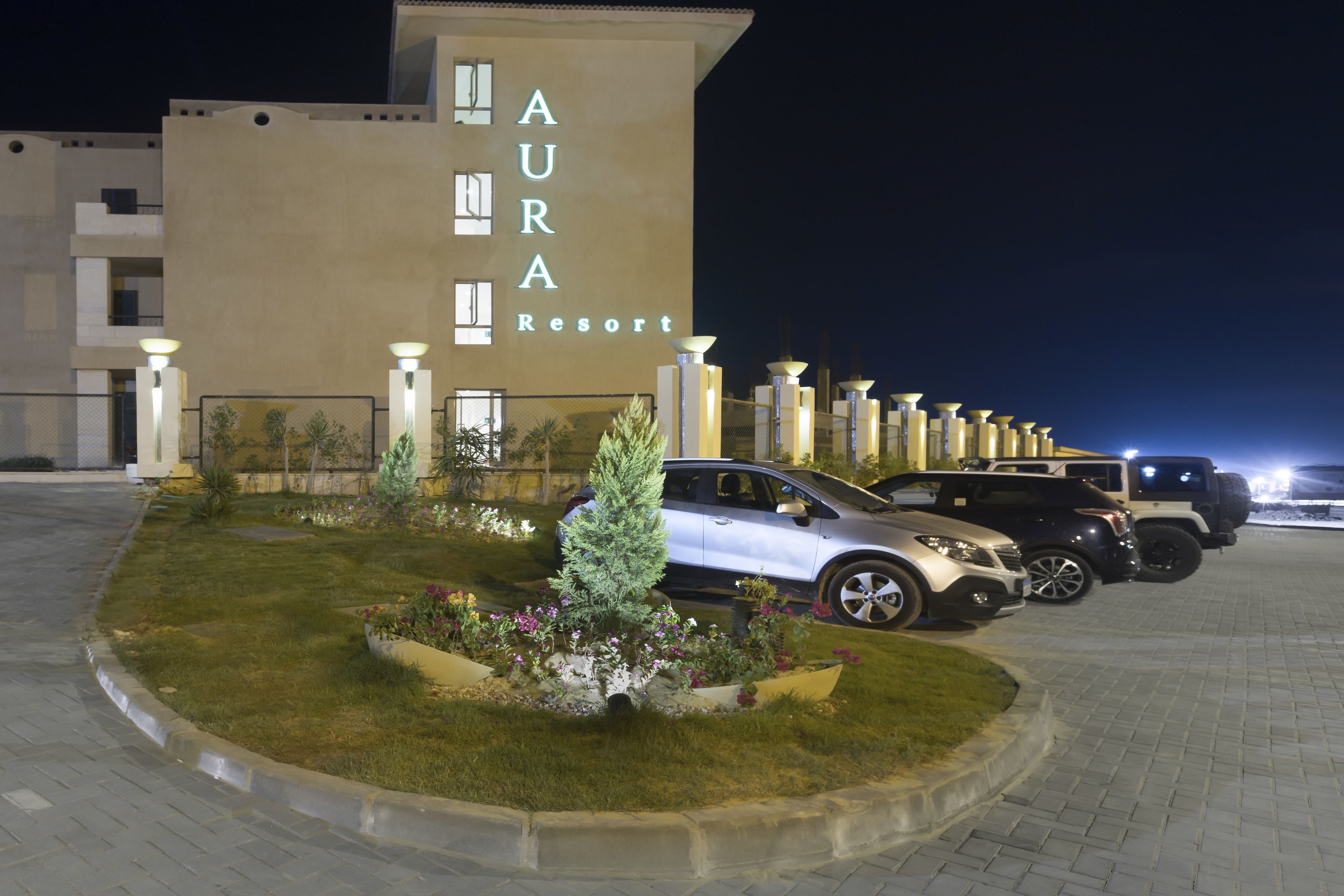 A hotel named "AURA Resort" with a paved driveway, parked cars, and a landscaped garden area.